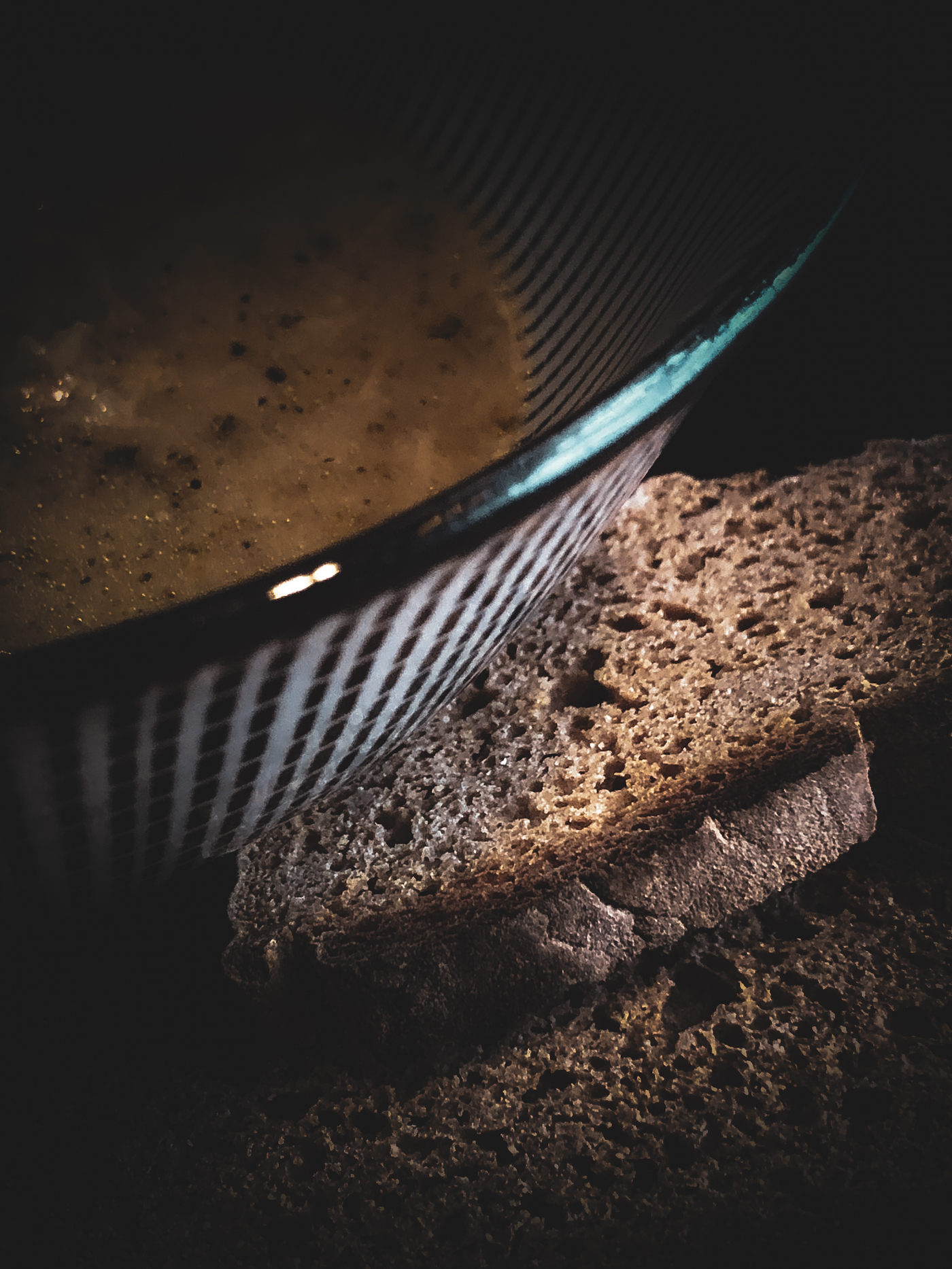 Pieces of rustic bread on a plate beside a bowl of soup, with light highlighting the bread’s texture in a moody setting.
