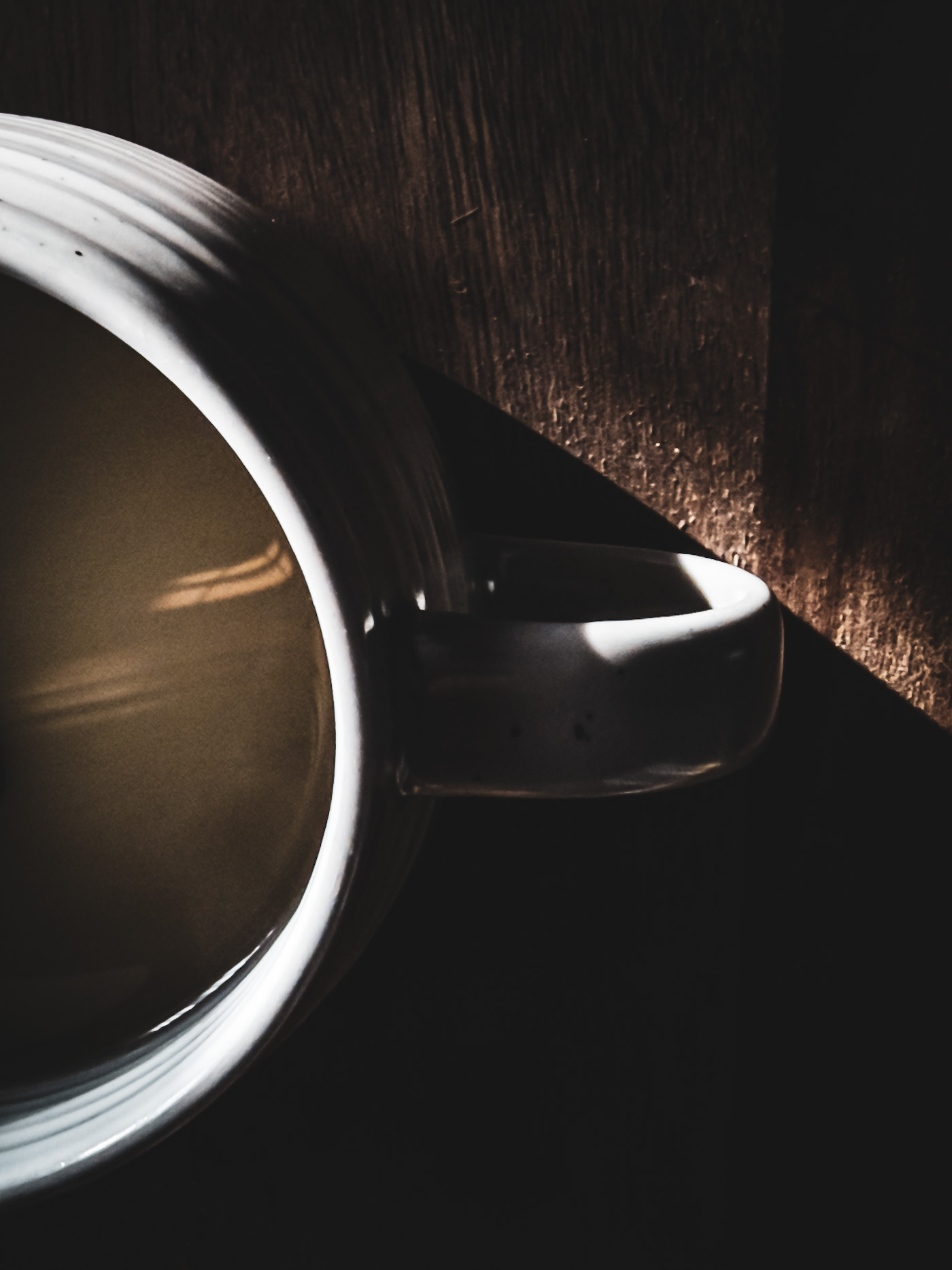 Top view of a mug of tea on a rustic wooden board, with strong light highlighting the rim and handle while the rest falls into shadow.