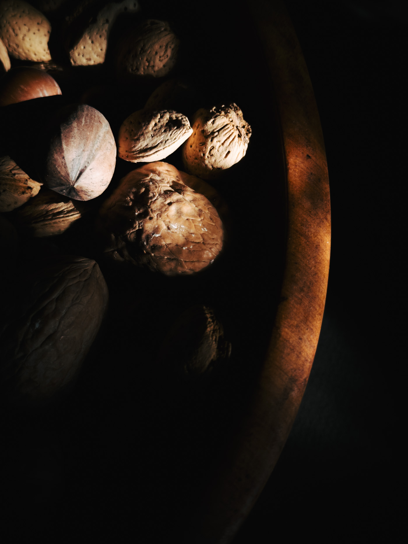 Top view of a wooden bowl filled with mixed nuts, softly revealed by light and partially hidden in shadow