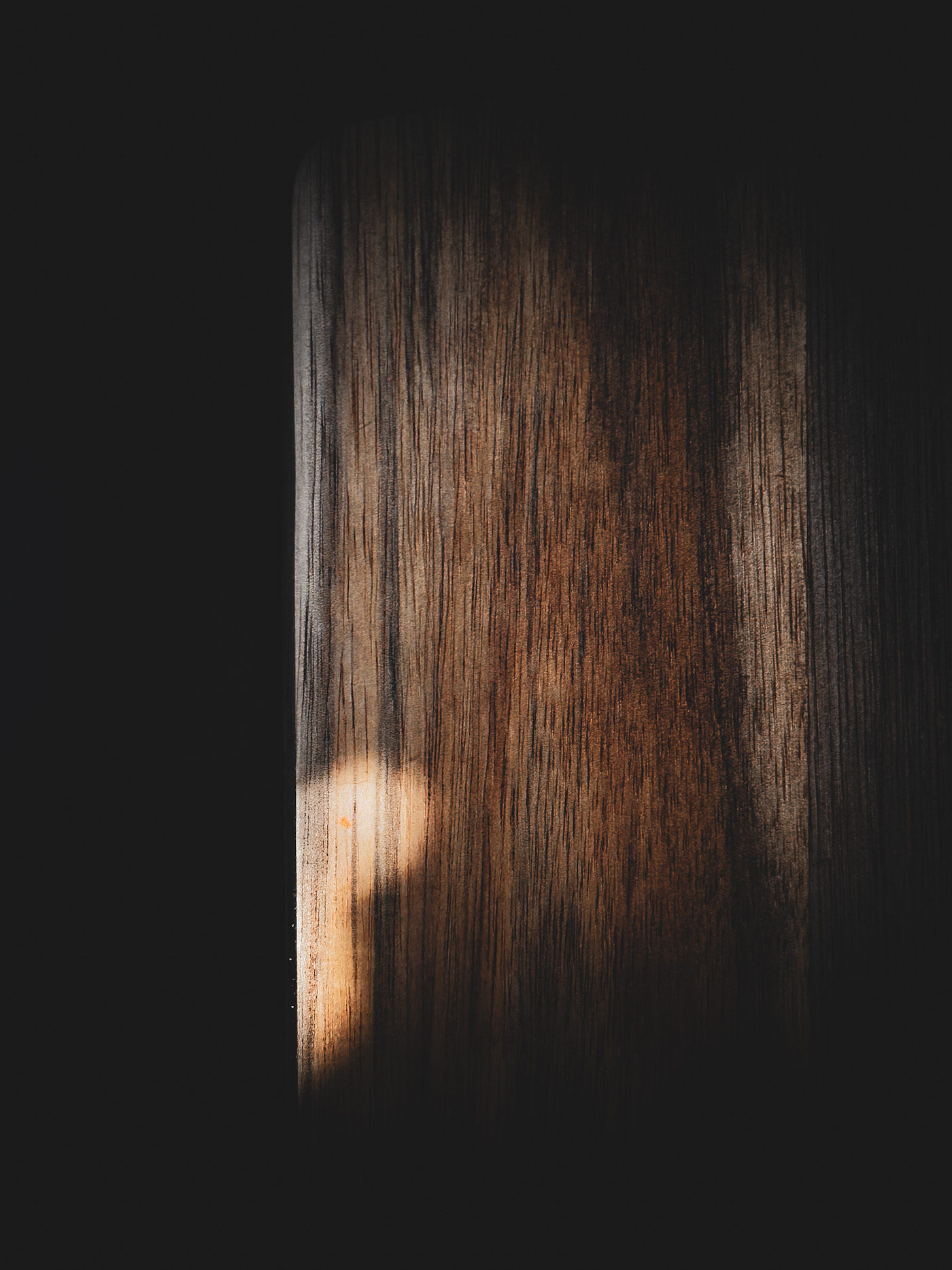 A quiet, atmospheric image of a wooden cutting board standing upright in deep shadow, catching a soft blob of light on one edge.